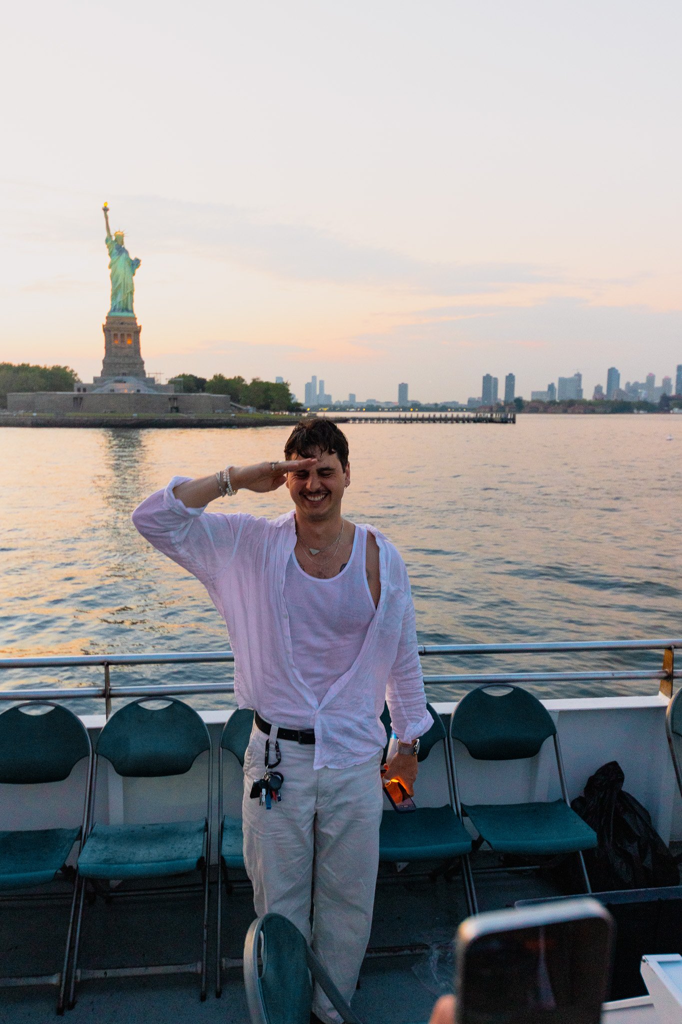 Adam Metwally smiling and saluting on a boat near the Statue of Liberty